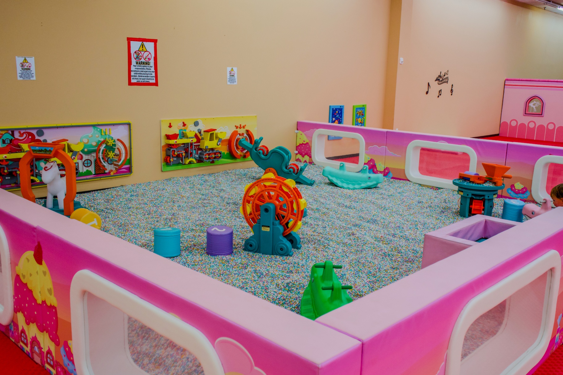 Colorful indoor sandbox play area with a large orange and blue toy wheel, whale rocker, mirrors and sensory toys surrounded by pink padded walls at Star Park