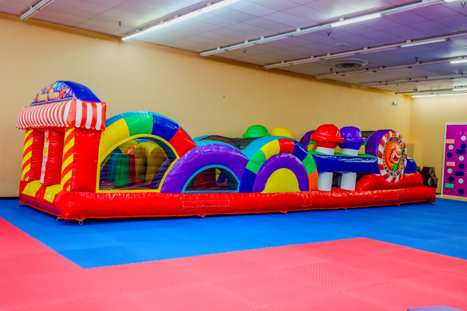 Colorful inflatable candy-themed obstacle course with red, purple, yellow and green arches, a candy cane striped entrance, and mushroom decorations on a blue and pink padded floor at Star Park