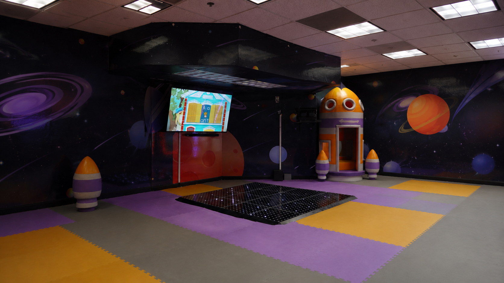 Space-themed indoor interactive play area with a glowing light-up floor, orange and purple rocket ship, rocket-shaped seats, planet murals and a large screen at Star Park