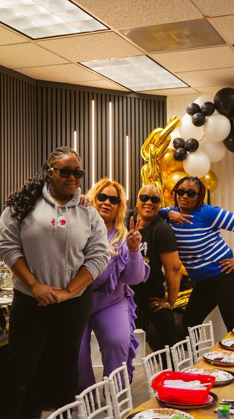 Four smiling women in sunglasses posing together with peace signs in a Star Park indoor playground party room decorated with a gold and black balloon arch, gold foil letter balloons and a long celebration table set with plates