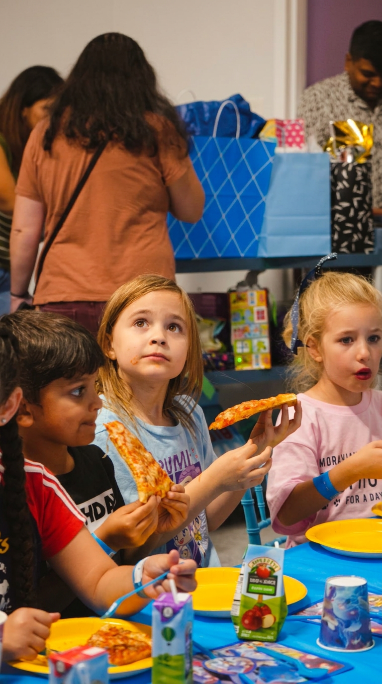 Three young girls eating pizza and drinking apple juice boxes at a Star Park birthday party table with blue tablecloth, yellow plates and blue gift bags in the background