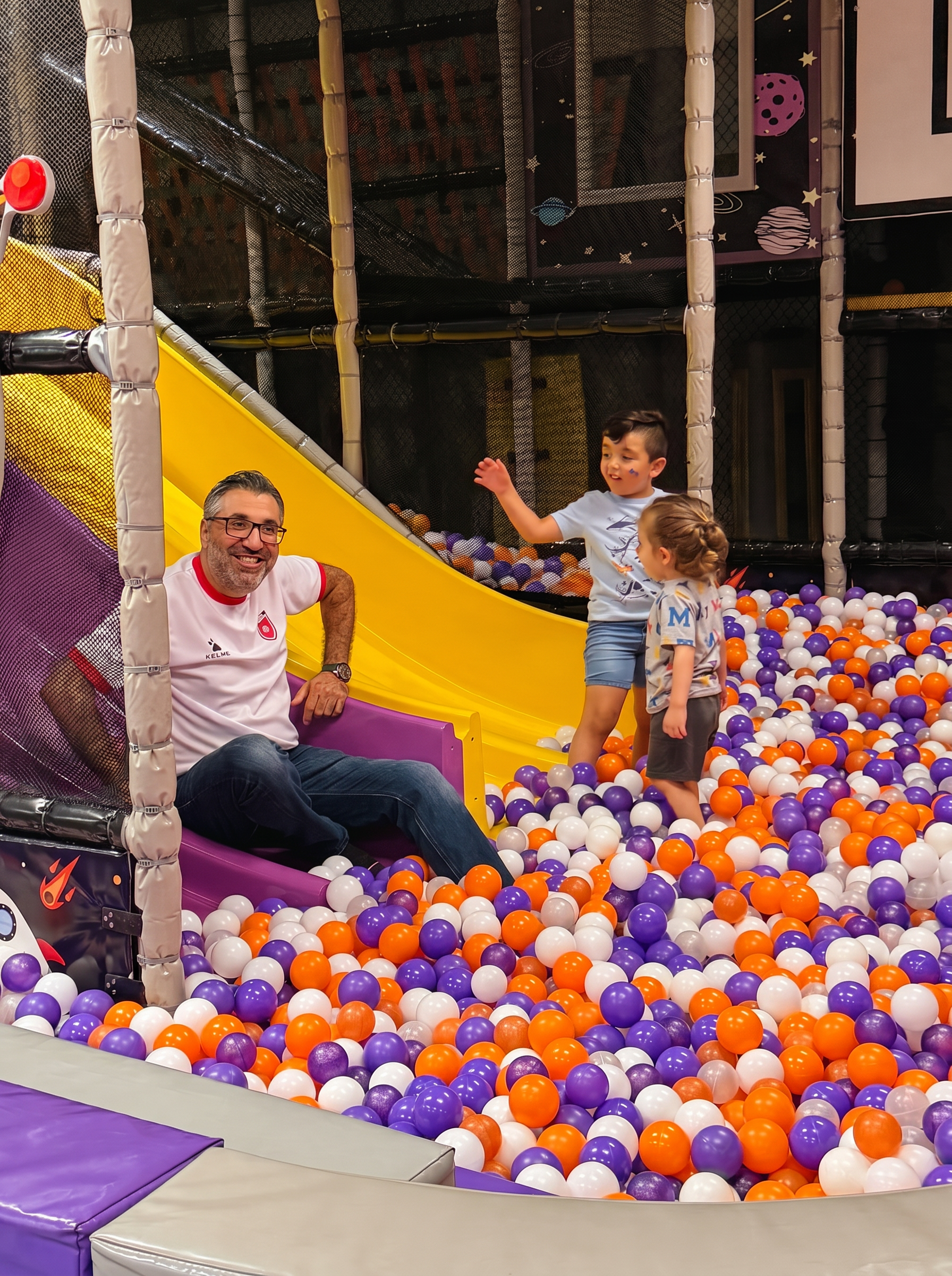 Smiling dad and two kids playing in the orange and purple ball pit beside the yellow slide at Star Park indoor playground